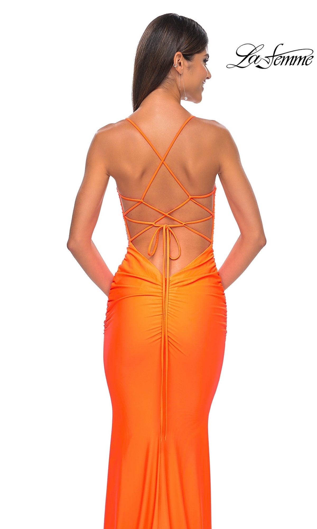 A woman stands with her back to the camera, wearing the eye-catching La Femme Backless Bright Long Prom Dress 32325. This floor-length orange gown features thin straps and a lace-up open back. The La Femme logo appears in the top right corner.